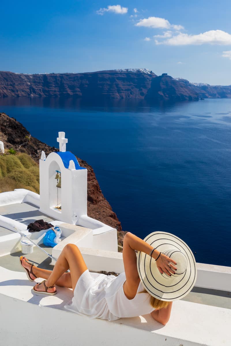 woman in white dress and white hat lounging on while building overlooking deep blue sea