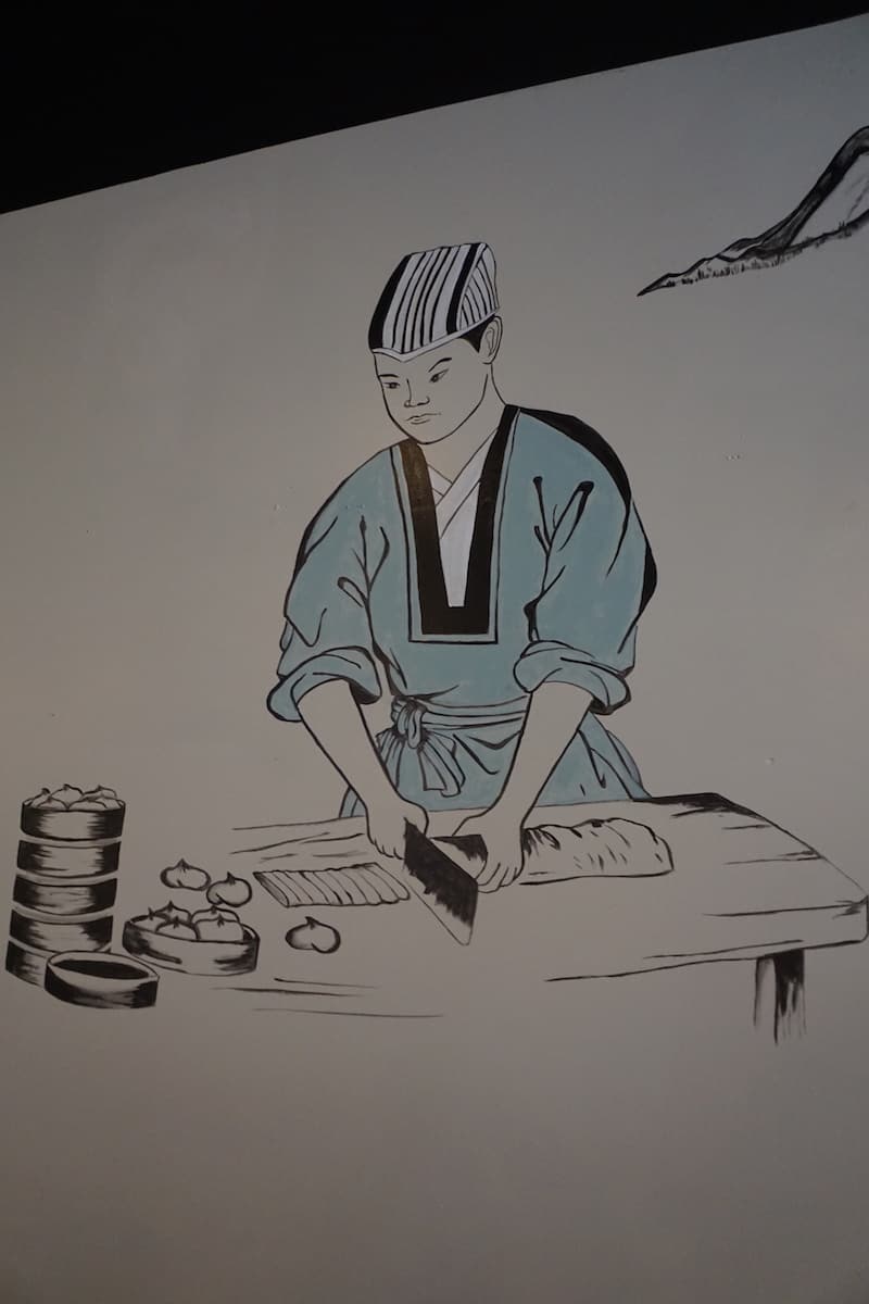 black and white mural of Japanese chef in teal coat making dumplings