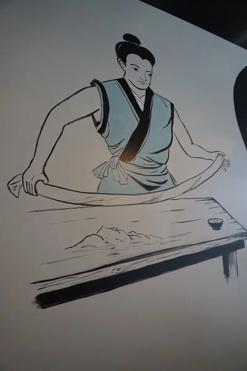 black and white mural of Japanese chef in teal coat making noodles by hand