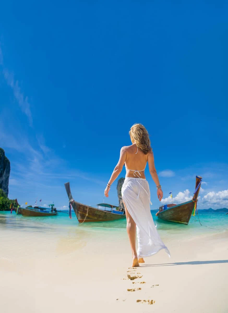 Young blonde woman wearing white bikini and white sarong walking on a white sand beach towards two Thai boats in the ocean