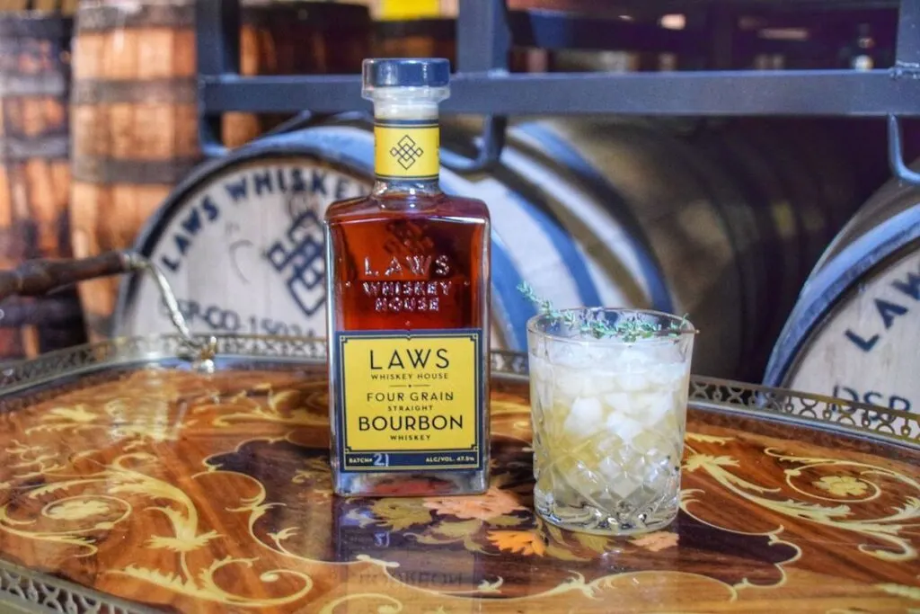 bottle of bourbon whiskey and pale yellow whiskey cocktail on brown decorative tray in front of bourbon barrels