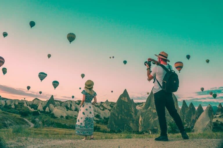 man taking photo of hot air balloons and blonde woman wearing maxi dress and straw hat in cappadocia
