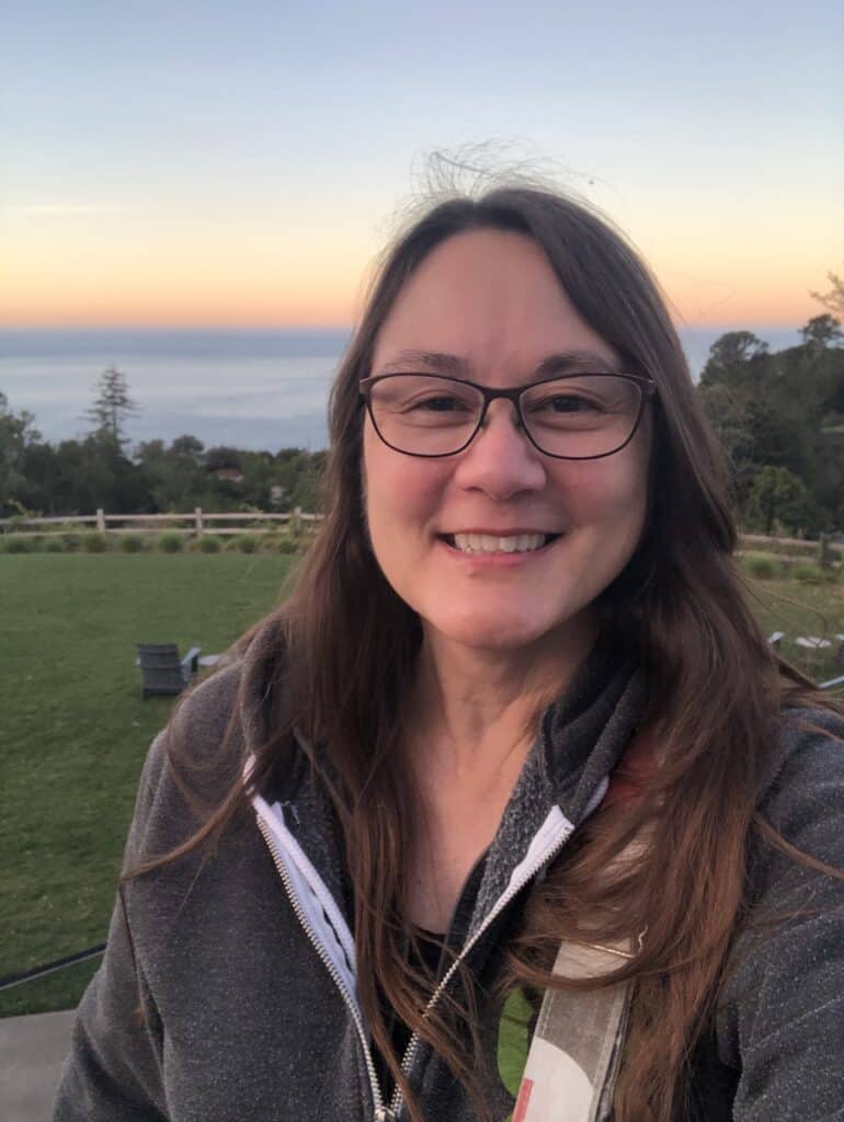 woman with brown hair wearing gray hoodie with green lawn and ocean in background at sunrise at Alila Ventana Big Sur