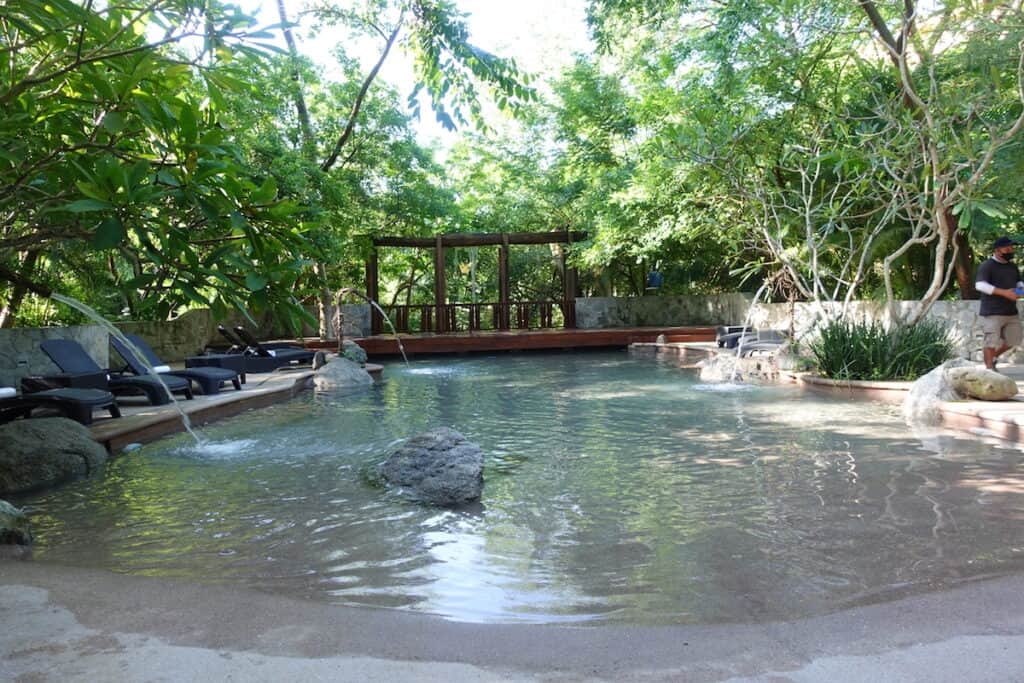 lake like walk in pool surrounded by green trees and black pool lounge chairs