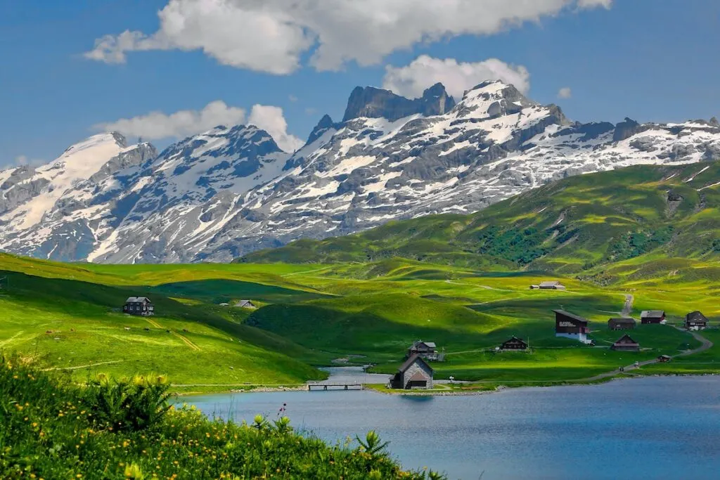 gray and black houses on vibrant green grass lands near lake in front of snow capped mountains, mountain and houses photo, switzerland