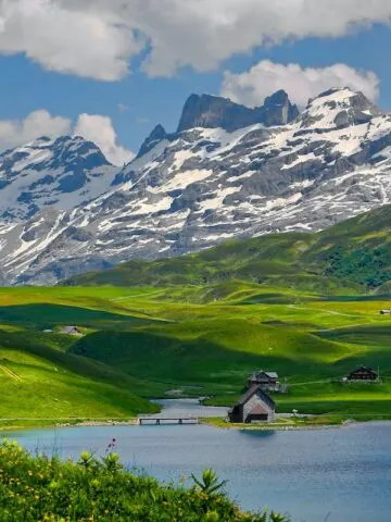 gray and black houses on vibrant green grass lands near lake in front of snow capped mountains, mountain and houses photo, switzerland