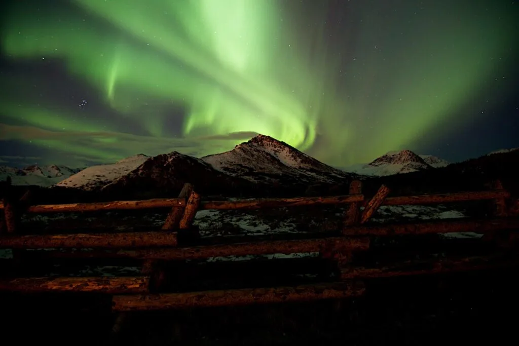 light green swirls among stars in dark night sky behind snow covered mountains behind brown log fence, Aurora Borealis illustration