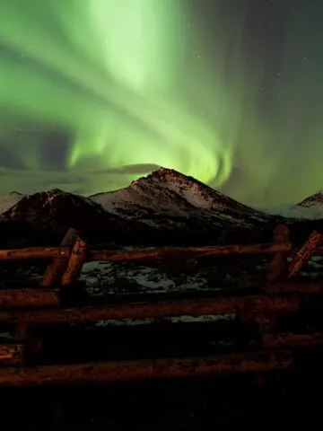 light green swirls among stars in dark night sky behind snow covered mountains behind brown log fence, Aurora Borealis illustration