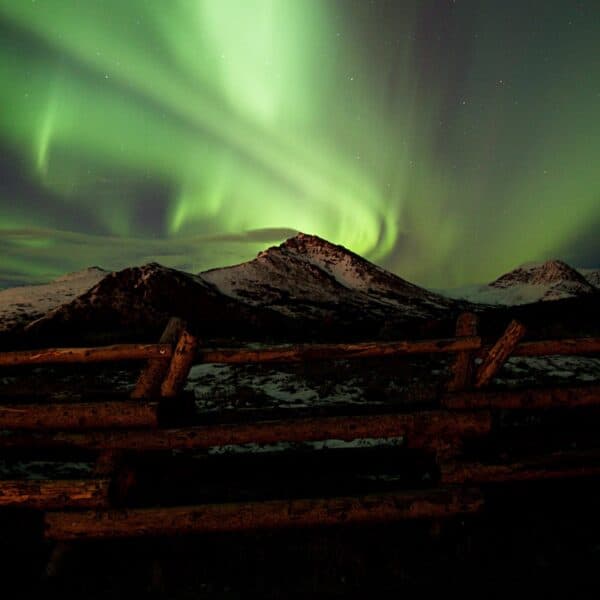 light green swirls among stars in dark night sky behind snow covered mountains behind brown log fence, Aurora Borealis illustration