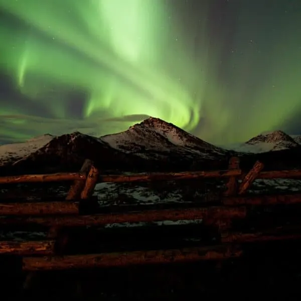 light green swirls among stars in dark night sky behind snow covered mountains behind brown log fence, Aurora Borealis illustration