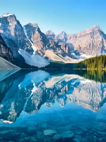 brown snow covered mountains and dark freen tall trees behind crystal clear mirror lake reflecting image on lake surface, scenery of mountain, moraine lake canada