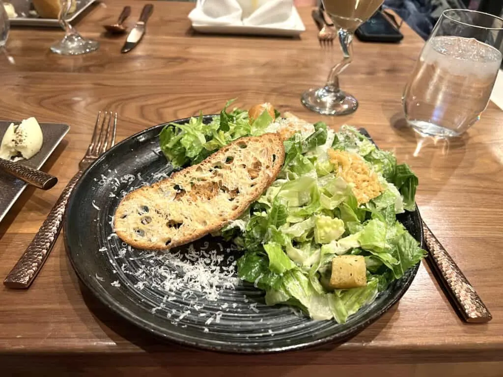 leafy greens topped with shredded parmesan on black plate and slice of toasted bread on brown table at restaurant