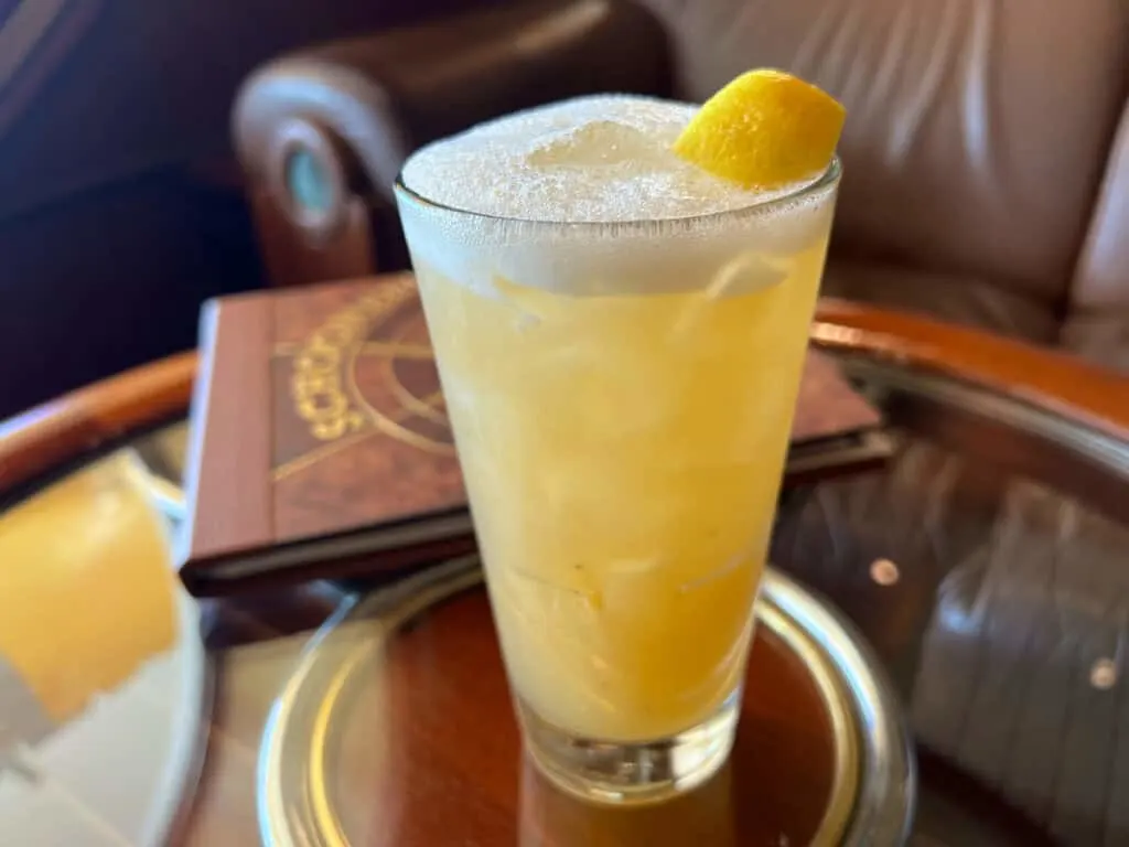 frothy yellow orange liquid in tall glass with lemon wedge on top of round glass table with wood edge