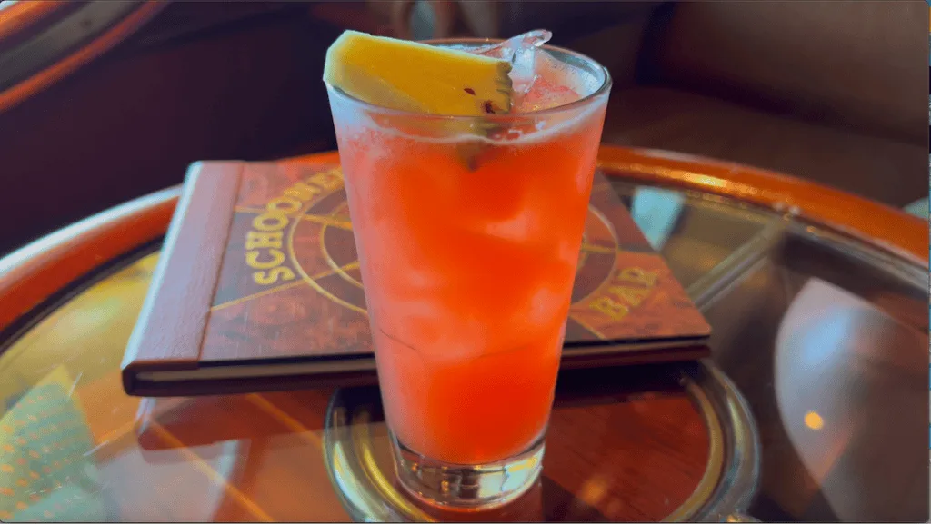 dark red orange liquid in tall glass topped with pineapple slice sitting on round glass table with wood edge