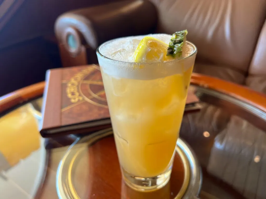 frothy orange brown liquid in tall glass topped with lemon wedge and pineapple slice sitting on round glass table with wood edge