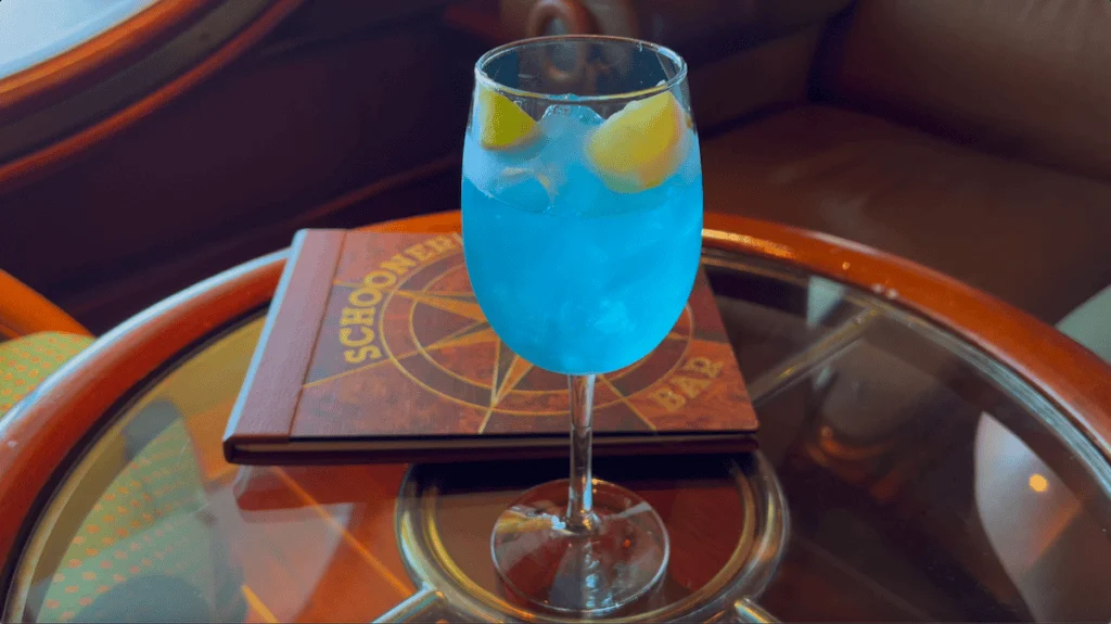 neon blue liquid in curved wine style glass with two lemon wedges sitting on round glass table with wood edge