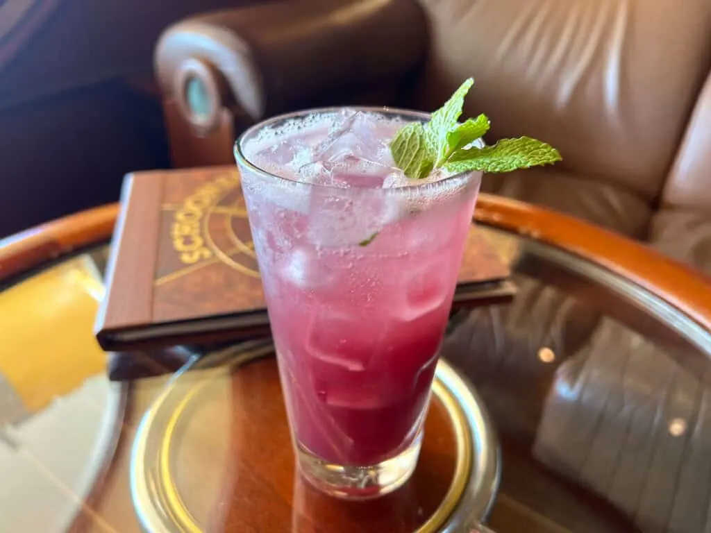 Purple liquid in tall glass with mint garnish on top of round glass table with wood edge