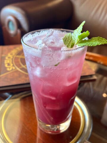 Purple liquid in tall glass with mint garnish on top of round glass table with wood edge