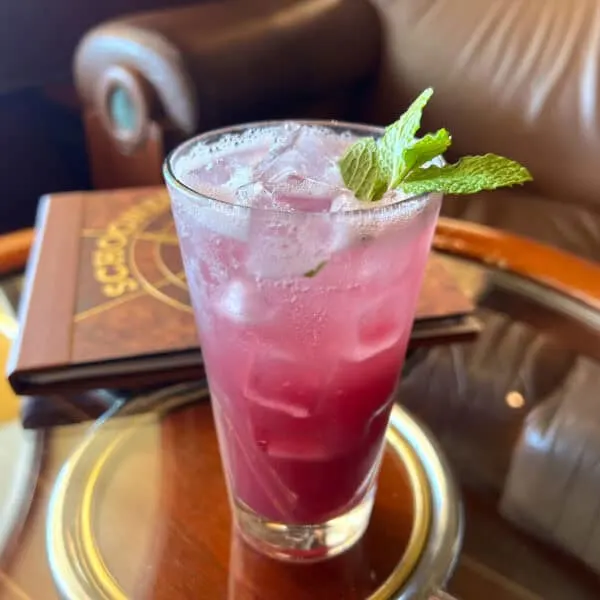 Purple liquid in tall glass with mint garnish on top of round glass table with wood edge