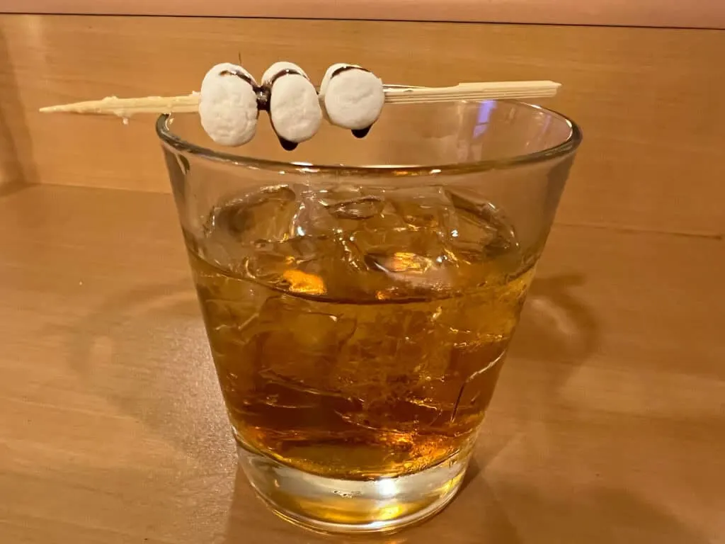wooden stick with white marshmallows and chocolate syrup on top of glass tumbler holding brown liquid alcohol 