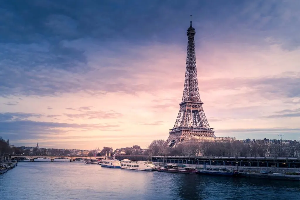 Tall skinny triangle shaped tower next to river with house boats in middle of city at dusk. Eiffel Tower, Paris France