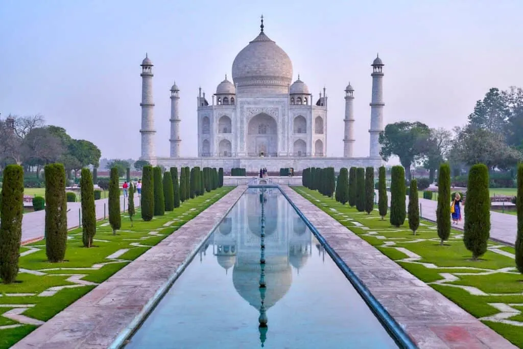 white gray building with Moorish towers and columns in front of narrow veritical pool flanked by tall dark green trees, photo of Taj Mahal, Entering the Taj Mahal at the sunrise, Agra, India