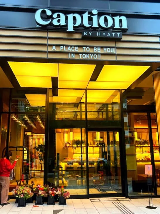 Black haired Asian woman wearing long sleeved red shirt and brown pants adjusts orange birds of paradise in front of a dark brown almost black hotel exterior with yellow cream lighting