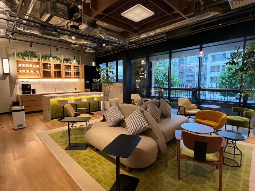 large community room with beige overstuffed sofas, orange chairs, green chairs, beige chairs, black tables, bar with stools, and outside terrace