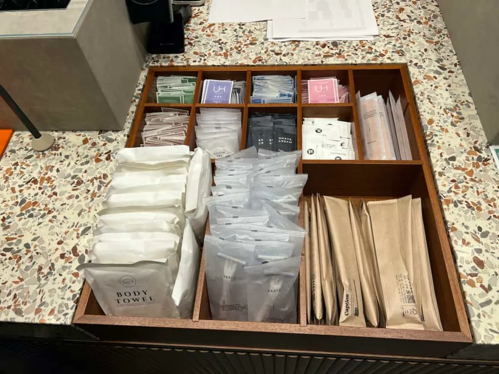 Wood tray with divided compartments holding various hotel amenties: individual body towels, razors, toothbrushes, toothpaste, combs, shaving cream