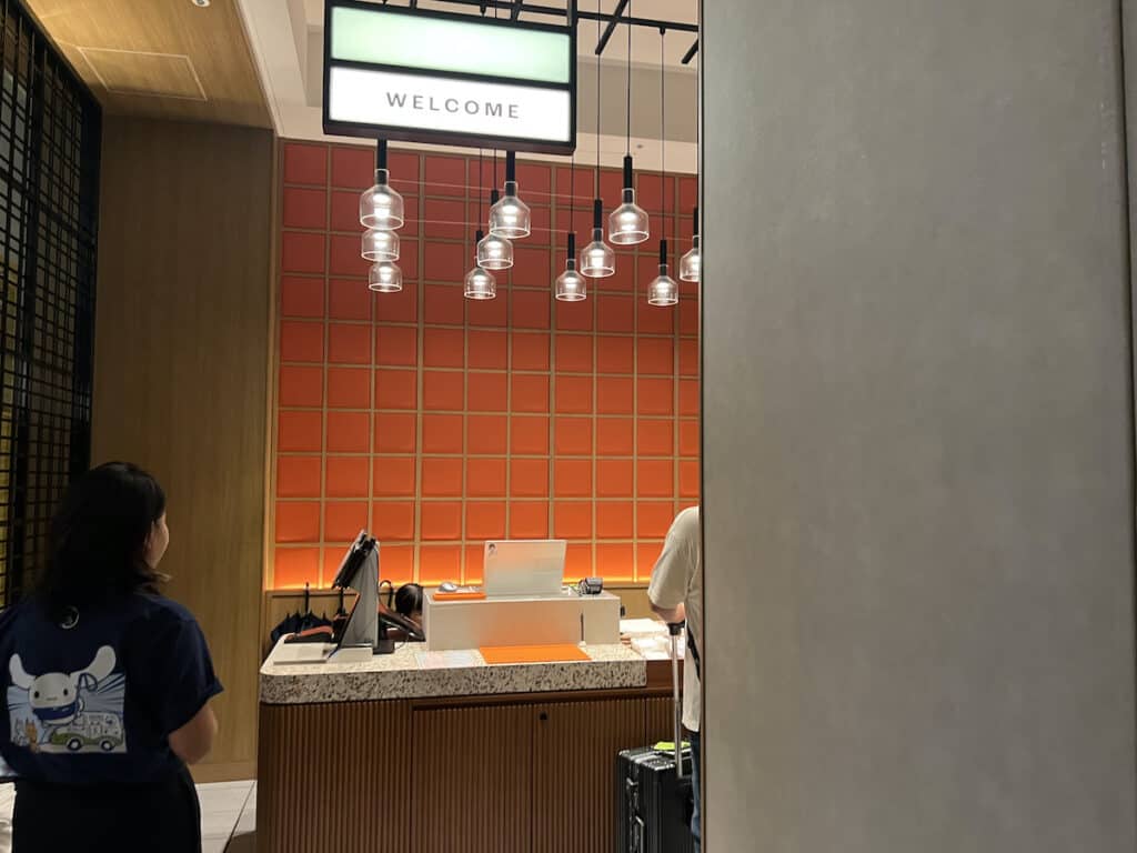 Black haired Asian woman wearing a black shirt with Asian cartoon character standing near quartz counter topped wood desk in front of a red tile background and multiple hanging lights at a hotel front desk