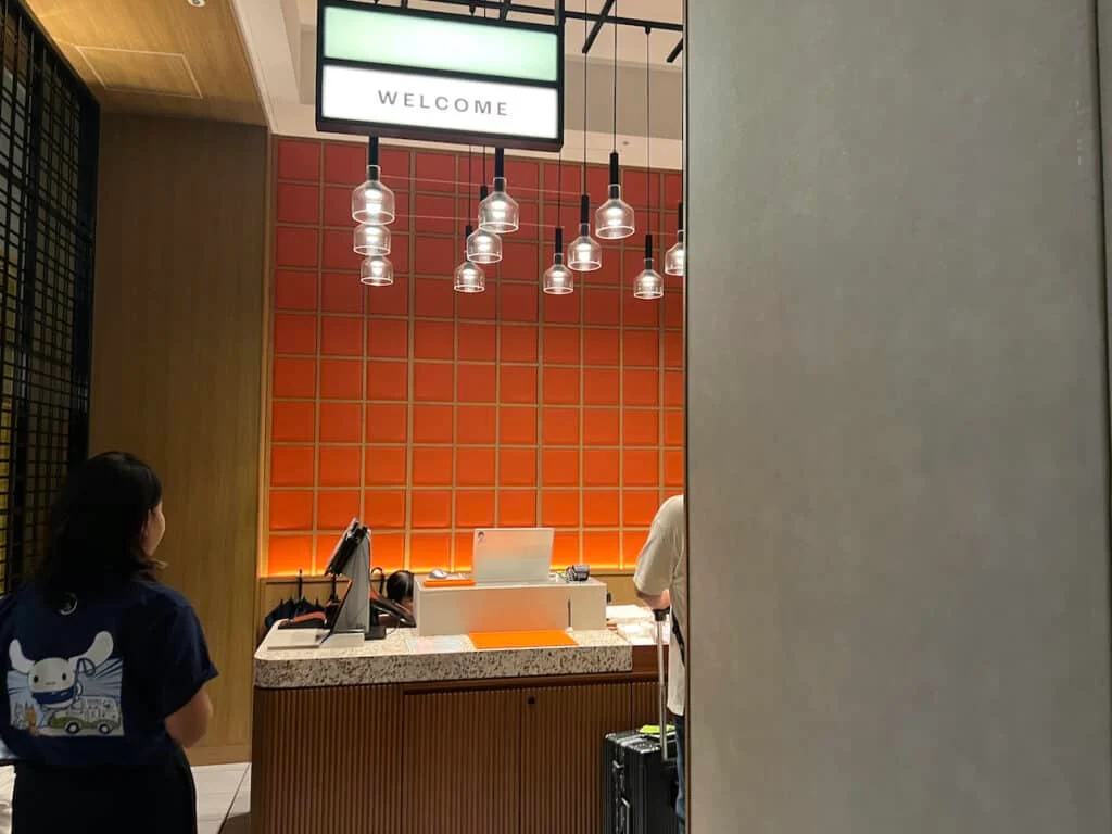 Black haired Asian woman wearing a black shirt with Asian cartoon character standing near quartz counter topped wood desk in front of a red tile background and multiple hanging lights at a hotel front desk