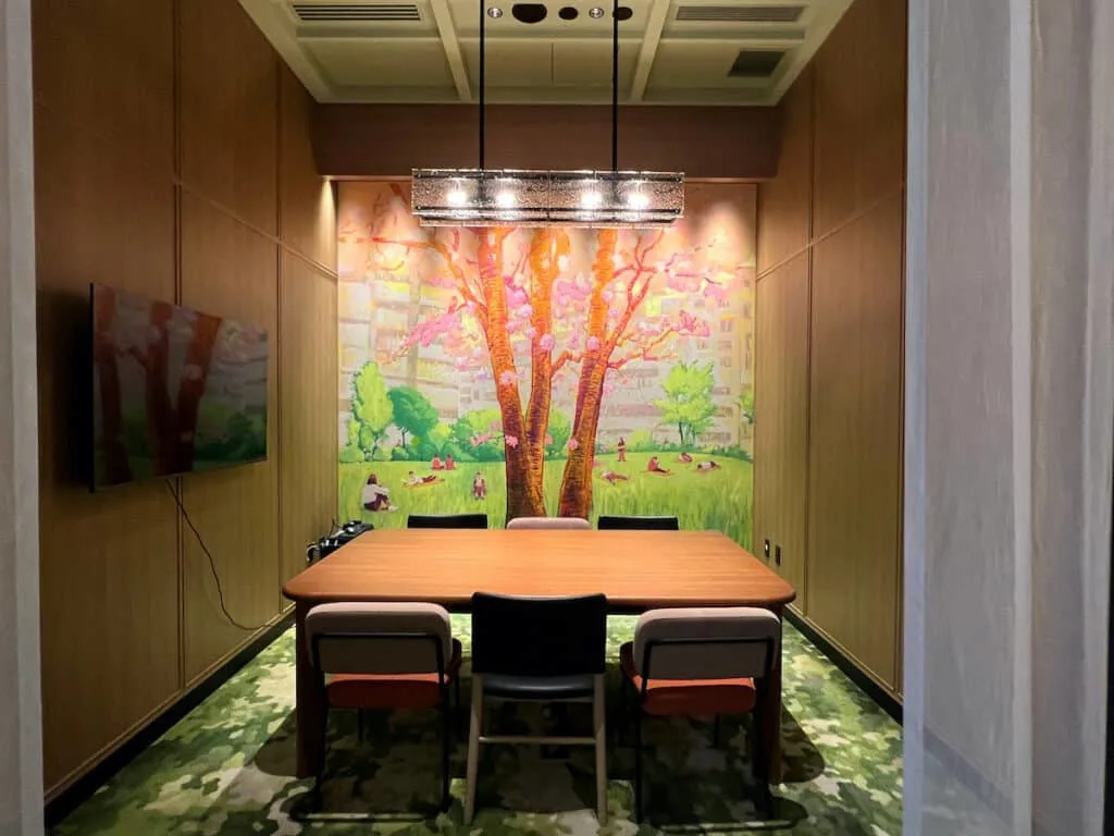 Six chairs around wood table in small meeting room with a flat screen TV on one wall and a mural of cherry blossom tree and people laying in grass with trees in park setting on wall facing viewer