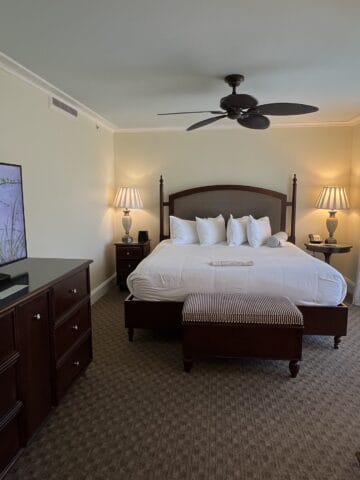 hotel room with brown wooden bed with white sheets, brown ceiling fan, flat screen tv on top of brown dresser