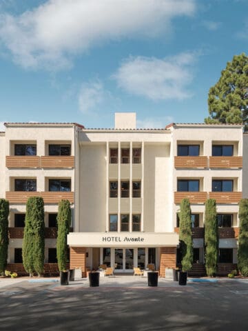 cream colored 4 story hotel with brown wood slat balconies and tall skinny green hedges in front of hotel