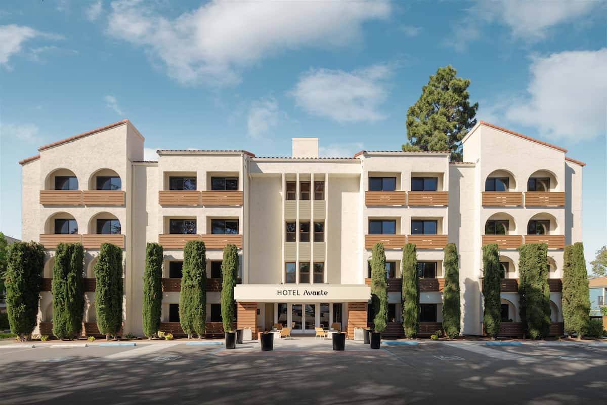 cream colored 4 story hotel with brown wood slat balconies and tall skinny green hedges in front of hotel