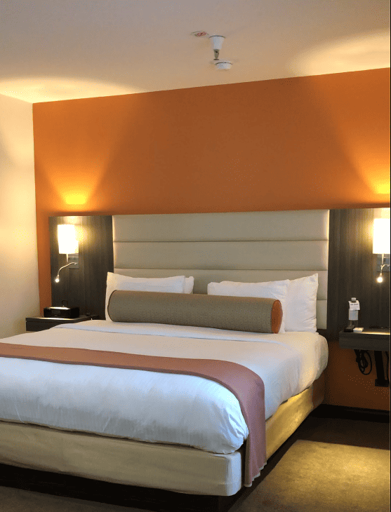 Bed with white linens, gray padded headboard against orange wall