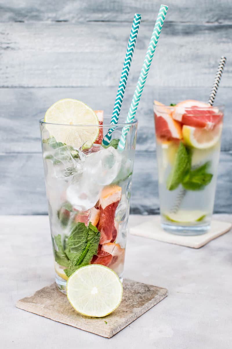 two clear glasses filled with water, fresh mint leaves, grapefruit, lime, and lemon slices and a paper straw