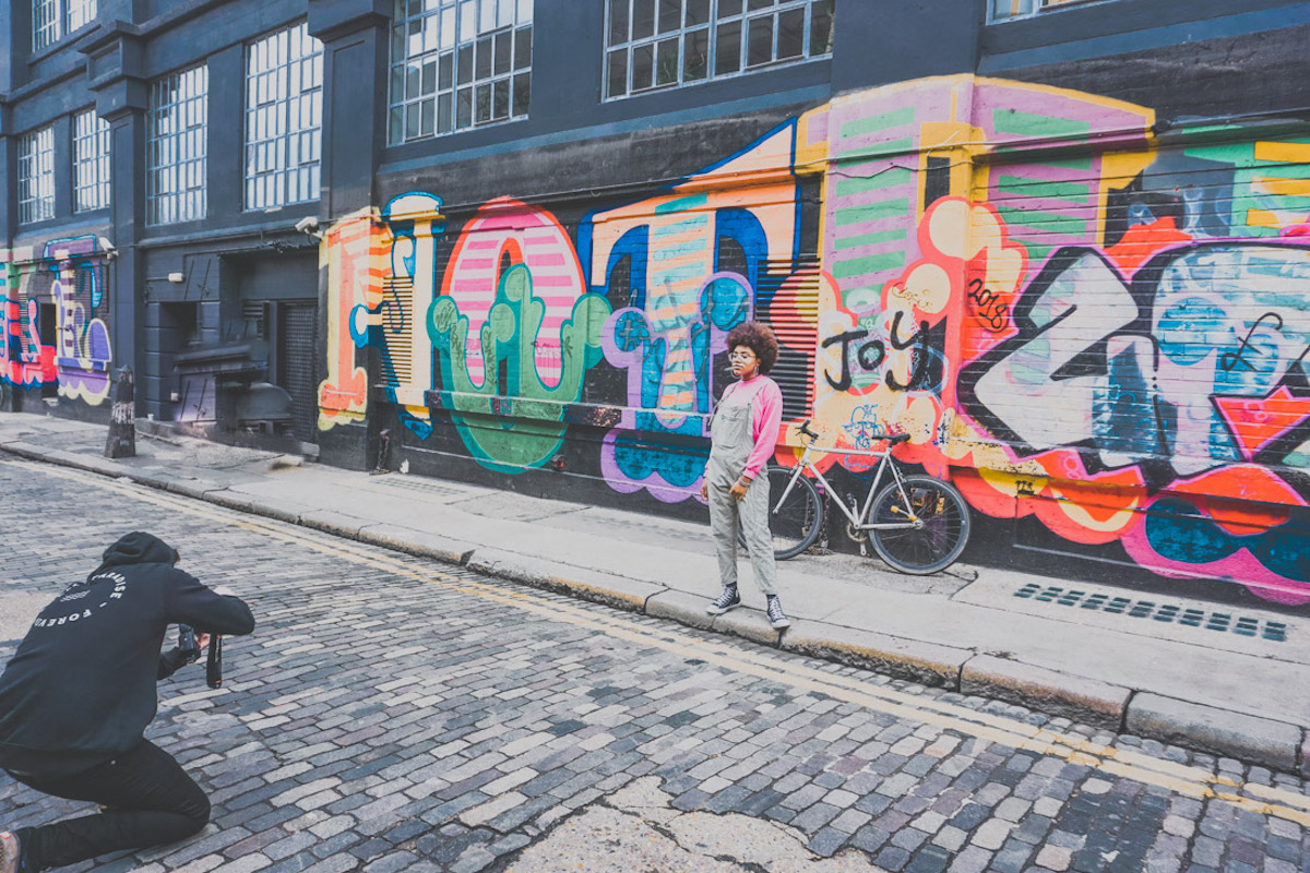 photographer dressed in black taking photo of girl in front of street art in Shoreditch London